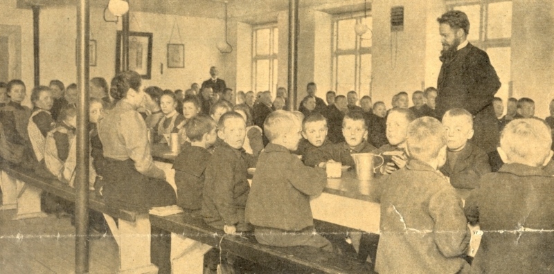 1907 Piger og drenge sidder hver for sig, når måltiderne skal indtages. Endnu i 1907 spiser og drikker man af tintallerkener og tinkrus. Lærerne sidder med til bords for at opretholde ro og orden.