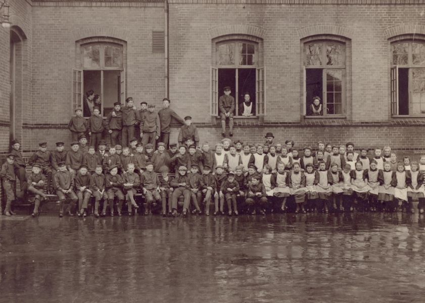 1909 Den nyudnævnte forstander, Anders Hansen, har samlet alle eleverne i den regnvåde gård. At en dreng har fået lov til at sidde i vindueskarmen, er vist ikke helt efter Anders Hansens regler.