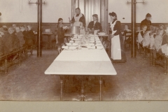 1907 Så har eleverne indtaget deres pladser (på hårde bænke) ved middagsbordet, piger og drenge hver for sig, naturligvis. Lærerne lader vente på sig, og det er køkkenpersonalet, i stilfuld påklædning, der betjener eleverne