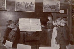 1907 Korlæsning efter Korlæsebog. Korlæsning i Fredericia med lærerinden som dirigent. Efter de ophængte anskuelsestavler at dømme, er emnet livet på landet